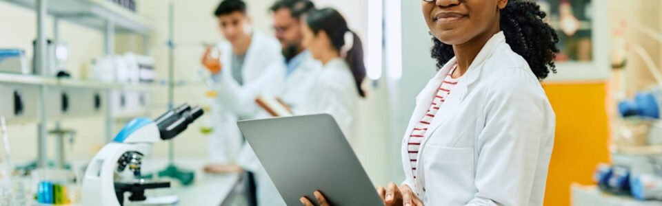 smiling woman in a modern lab, uses pharma contract management on her laptop